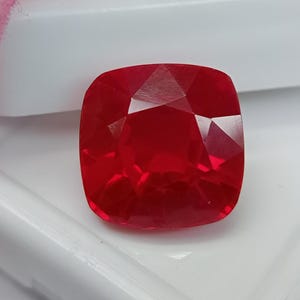 Puede incluir: Una gema roja vibrante de corte cojín con una superficie facetada que refleja la luz. La piedra muestra un tono carmesí profundo y saturado y una forma cuadrada redondeada simétrica, con cortes geométricos precisos en su superficie.