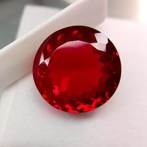 Könnte beinhalten: Ein runder, facettierter Edelstein in einer tiefen, leuchtenden roten Farbe. Der Stein weist einen Brillantschliff mit zahlreichen dreieckigen und trapezförmigen Facetten auf, die das Licht reflektieren und eine klare, polierte Oberfläche erzeugen.