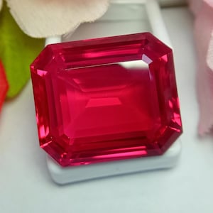May include: A vibrant, deep red gemstone with an emerald cut, featuring sharp, clean facets that reflect light across its rectangular surface. The stone displays a uniform, saturated crimson hue throughout its transparent, polished crystalline structure.