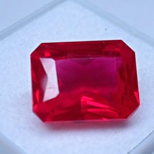 Könnte beinhalten: Ein leuchtender, rechteckig geschliffener Edelstein in einer tiefen, gesättigten roten Farbe. Der Stein weist zahlreiche Facetten auf, die scharfe, geometrische Reflexionen erzeugen und eine hohe Klarheit für ein brillantes, transparentes Aussehen.