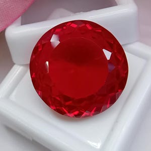 Könnte beinhalten: Ein runder, facettierter, leuchtend roter Edelstein im Brillantschliff mit zahlreichen dreieckigen und rautenförmigen Facetten, die das Licht reflektieren. Der Edelstein ist mittig auf einer weißen, quadratischen Schmuckschatulle platziert.