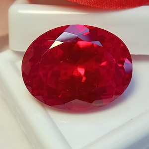 Könnte beinhalten: Ein leuchtender, oval geschliffener Edelstein mit einer tiefen, gesättigten roten Farbe und zahlreichen Facetten, die eine brillante, reflektierende Oberfläche erzeugen. Der Stein liegt auf einer glatten, weißen, geschwungenen Basis.