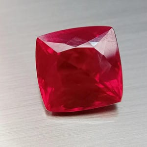 May include: A vibrant, deep red gemstone with a cushion cut, featuring multiple triangular and trapezoidal facets that create a reflective, brilliant surface. The stone has a square-like shape with slightly rounded corners and a translucent quality.