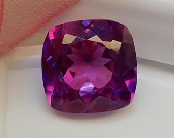 May include: A cushion-cut gemstone with a vibrant purple and magenta color gradient. The stone features a faceted surface that creates a star-like pattern in the center, reflecting light with high clarity and a polished, glossy finish.