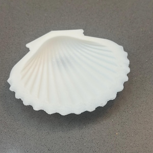 Könnte beinhalten: Eine weiße, muschelförmige Muschel mit strukturierter Oberfläche. Die Muschel hat ein geripptes Muster, das vom Zentrum ausgeht, und einen gewellten Rand. Die Muschel liegt auf einer grauen Oberfläche.