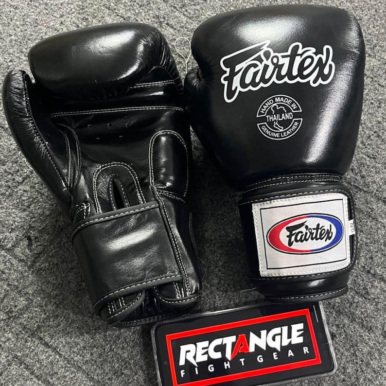 May include: Pair of black Fairtex boxing gloves with white stitching. The gloves have the Fairtex logo in white and a label that says "Hand Made in Thailand Genuine Leather". A Rectangle Fight Gear logo is visible.