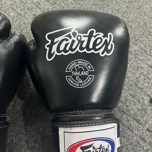 May include: Black Fairtex boxing gloves with white logo and text. The gloves have a white label that says "Hand Made in Thailand Genuine Leather". The gloves are designed for combat sports.