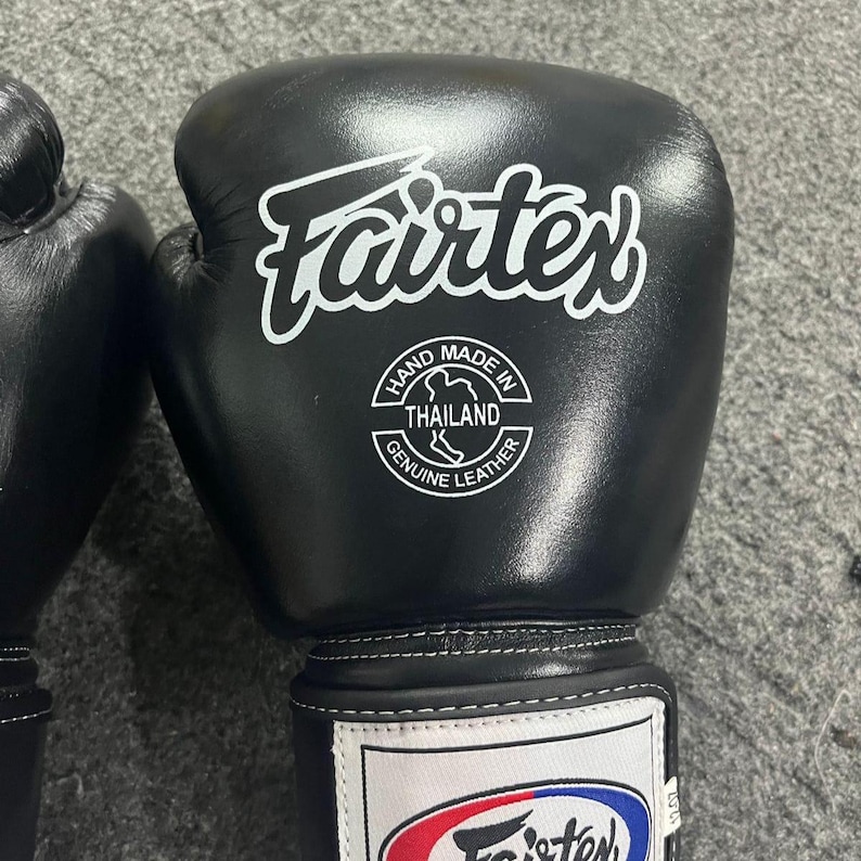 May include: Black Fairtex boxing gloves with white logo and text. The gloves have a white label that says "Hand Made in Thailand Genuine Leather". The gloves are designed for combat sports.