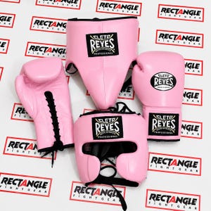 Peut inclure: Un ensemble d'équipement de boxe rose comprenant une paire de gants de boxe à lacets, un casque avec un écusson noir et une coquille de protection. Chaque article porte le logo Cleto Reyes en noir et blanc, avec des lacets et bordures noirs.