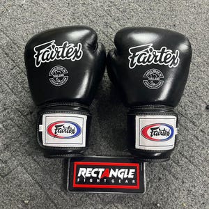 May include: Pair of black Fairtex boxing gloves with white logos and text. The gloves have a wrist strap with the Fairtex logo in red, white, and blue. The gloves are labeled "Hand Made in Thailand Genuine Leather."