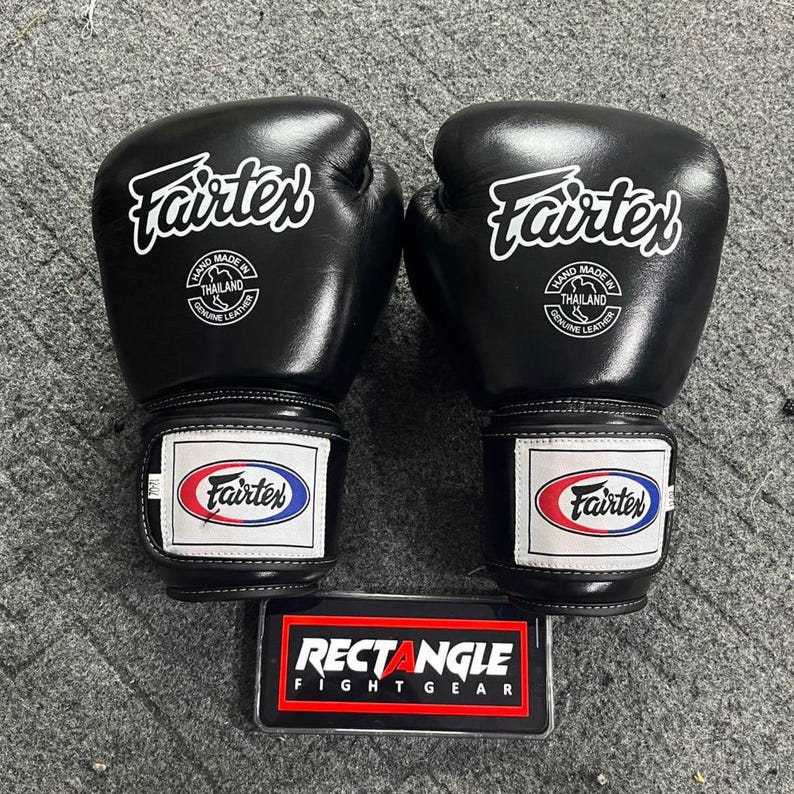 May include: Pair of black Fairtex boxing gloves with white logos and text. The gloves have a wrist strap with the Fairtex logo in red, white, and blue. The gloves are labeled "Hand Made in Thailand Genuine Leather."