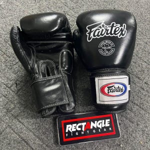 May include: Pair of black Fairtex boxing gloves with white logos and stitching. The gloves are made of genuine leather and feature a wrist strap with the Fairtex logo in red, white, and blue. A Rectangle Fight Gear logo is visible.