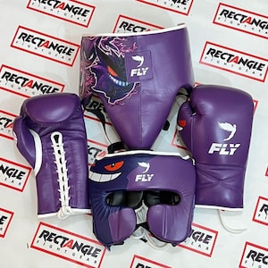 Puede incluir: Un conjunto de equipo de boxeo morado, que incluye guantes y protector de cabeza, con detalles blancos y el logotipo "FLY". El protector de cabeza presenta un gráfico de dibujos animados. Los guantes de boxeo tienen cordones blancos. El texto "RECTANGLE FIGHT GEAR" es visible en el fondo.