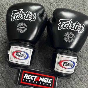 May include: A pair of black Fairtex boxing gloves with white logos and text. The gloves have a rectangular patch with the Fairtex logo in red, white, and blue. The text "RECTANGLE FIGHT GEAR" is visible.