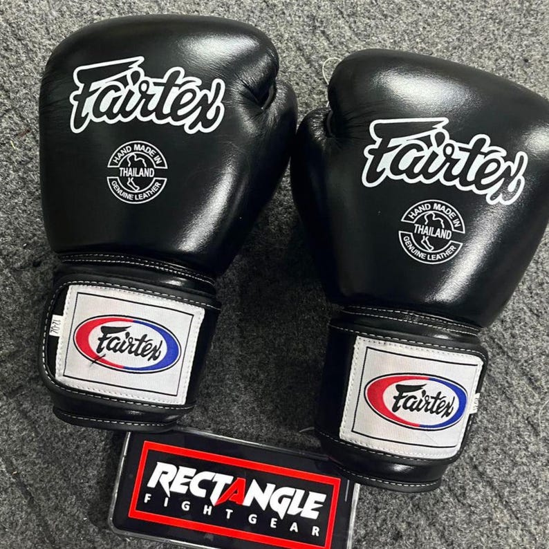 May include: A pair of black Fairtex boxing gloves with white logos and text. The gloves have a rectangular patch with the Fairtex logo in red, white, and blue. The text "RECTANGLE FIGHT GEAR" is visible.