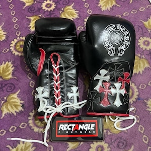 以下が含まれることがあります： 白と赤のアクセントが付いた黒いボクシンググローブ。十字のデザインと「Chrome Hearts」の文字が特徴です。グローブは白い紐で結ばれており、「RECTANGLE FIGHT GEAR」の文字が入った電話の隣にあります。