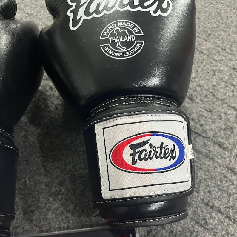 May include: Black Fairtex boxing gloves with white stitching. The gloves have a white patch with the Fairtex logo in red and blue. The gloves are labeled "Hand Made in Thailand Genuine Leather".