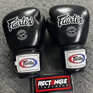 May include: A pair of black Fairtex boxing gloves. Each glove features the Fairtex logo in white, a patch with the Fairtex logo in red, white, and blue, and the text "HAND MADE IN THAILAND GENUINE LEATHER". The gloves are on a gray surface.