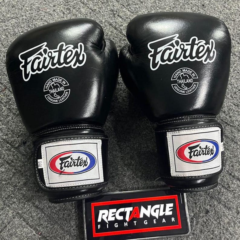 May include: A pair of black Fairtex boxing gloves. Each glove features the Fairtex logo in white, a patch with the Fairtex logo in red, white, and blue, and the text "HAND MADE IN THAILAND GENUINE LEATHER". The gloves are on a gray surface.