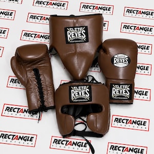 Peut inclure: Un ensemble d'équipement de boxe marron comprenant une paire de gants de boxe à lacets, un casque avec mentonnière et une coquille de protection. Chaque pièce porte une étiquette Cleto Reyes noire et blanche avec le texte Hecho en Mexico Professional.