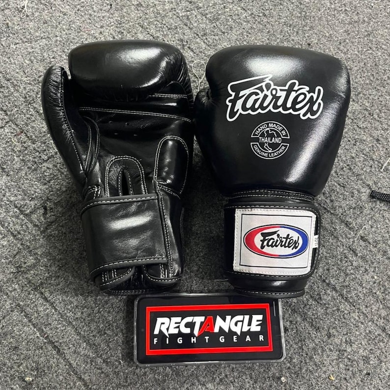 May include: A pair of black Fairtex boxing gloves. The gloves have white logos and text, including "Hand Made in Thailand Genuine Leather." A rectangular sign with the text "RECTANGLE FIGHT GEAR" is below the gloves.