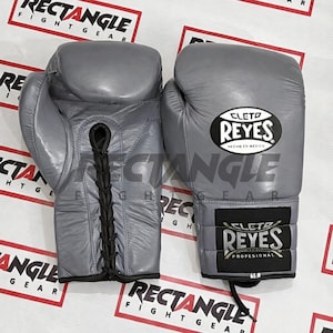Peut inclure: Une paire de gants de boxe en cuir gris avec des lacets noirs. Les gants portent le logo "CLETO REYES" en blanc sur un ovale noir et une étiquette rectangulaire noire avec le logo "REYES PROFESIONAL". Les gants sont marqués "16".