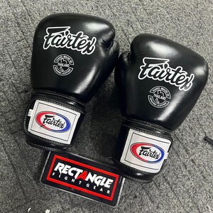 May include: A pair of black Fairtex boxing gloves with white logos and text. The gloves have a white and red patch on the wrist with the Fairtex logo. A black and red Rectangle Fightgear sign is below the gloves.