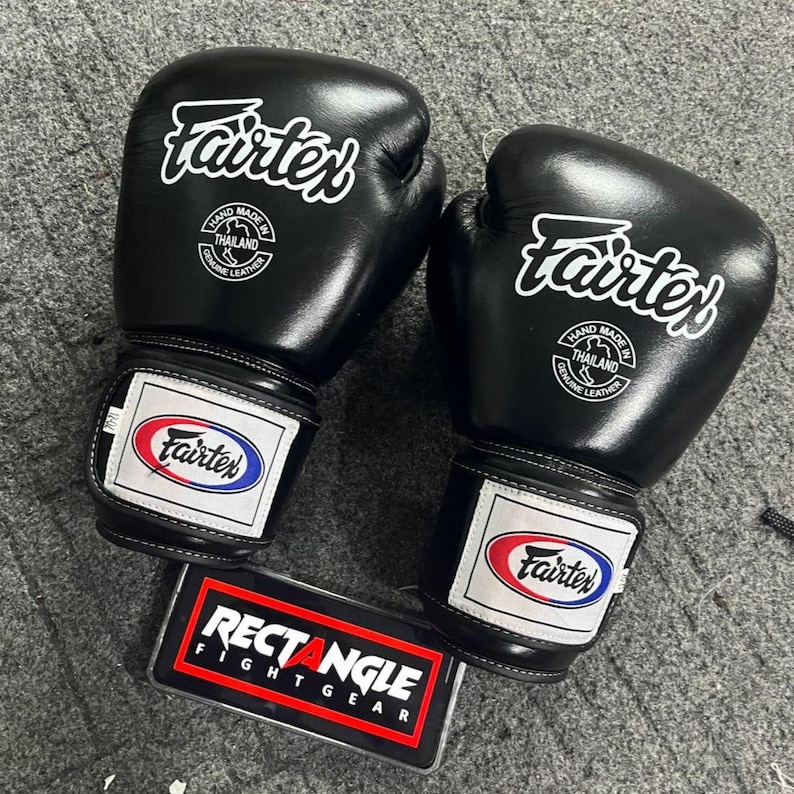 May include: A pair of black Fairtex boxing gloves with white logos and text. The gloves have a white and red patch on the wrist with the Fairtex logo. A black and red Rectangle Fightgear sign is below the gloves.