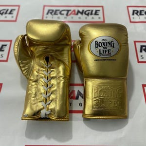Könnte beinhalten: Ein Paar goldene Boxhandschuhe. Einer der Handschuhe hat weiße Schnürsenkel und der andere das Logo "NO BOXING NO LIFE" und "HECHO EN MEXICO". Die Handschuhe sind aus glänzendem Material.