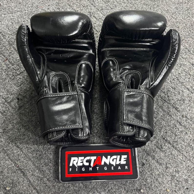 May include: A pair of black boxing gloves with white stitching. The gloves are made of a shiny material and have a hook-and-loop closure. A rectangular sign with the text "RECTANGLE FIGHT GEAR" is at the bottom.