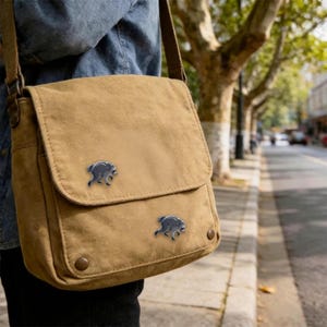 May include: A tan canvas messenger bag featuring two small, silver-toned metal enamel pins shaped like running raccoons attached to the front flap. The bag has a wide shoulder strap and two circular metal snap closures on the lower front corners.