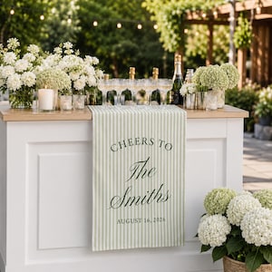 May include: A light green and white striped table runner draped over a white bar, featuring the dark green printed text "CHEERS TO The Smiths AUGUST 16, 2026". The runner is accented by white floral arrangements and clear glassware on the wooden surface.