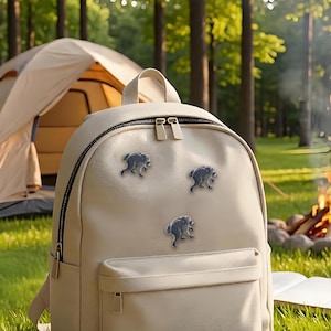 May include: A beige backpack featuring three small, metallic, silver-toned raccoon-shaped enamel pins attached to the front panel. The backpack has a main zippered compartment, a smaller front pocket, and a top handle with matching beige zippers.