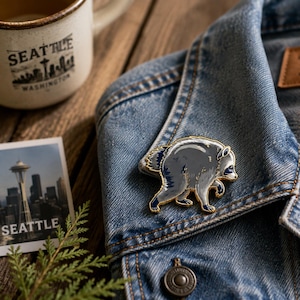 May include: A metal enamel pin shaped like a raccoon in a hunched, walking pose. The raccoon is colored in shades of grey, white, and dark blue with gold-toned metal outlines. The pin is attached to the collar of a blue denim jacket.