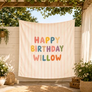 May include: A rectangular fabric backdrop featuring a cream background with vertical light pink stripes. The text "HAPPY BIRTHDAY WILLOW" is printed in the center using multi-colored, patterned bubble letters in red, blue, yellow, and green.
