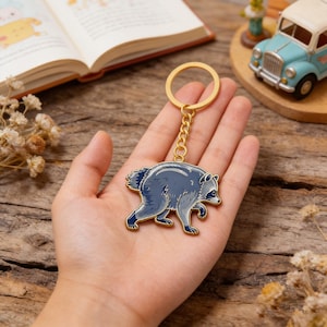 May include: A gold-toned metal keychain featuring a detailed enamel charm of a walking raccoon. The raccoon is depicted in shades of dark blue and grey with a black mask around its eyes. The charm is attached to a circular gold ring by a short chain.