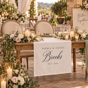 May include: A wooden sweetheart table featuring a white fabric runner printed with the text SOPHIA & DANIEL Brooks EST. 2026. The table is decorated with white and pink floral arrangements, eucalyptus greenery, and glass pillar candles.