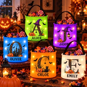 May include: Five cylindrical Halloween candy buckets with black handles, each featuring a large letter, a name, and LED lights. The buckets are lime green, purple, blue, orange, and white, decorated with pumpkins, bats, and spiderwebs.