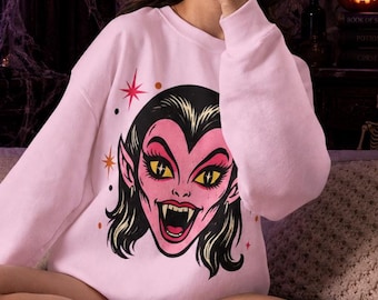 May include: A light pink long-sleeve sweatshirt featuring a graphic illustration of a vampire face with yellow eyes, sharp fangs, and black hair. The design includes small red and orange star accents on the sides of the vampire character's head.