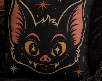 May include: A square throw pillow featuring a black background with a central graphic of a stylized bat face. The bat has large yellow eyes with star-shaped pupils, pink inner ears, and small white fangs. Orange and white star accents surround the bat.