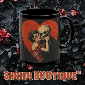 May include: Black ceramic mug featuring a romantic illustration of a couple kissing, with one partner a skeleton. The design is set against a red heart backdrop. The mug has the text "SHRIEK BOOTIQUE" at the bottom.