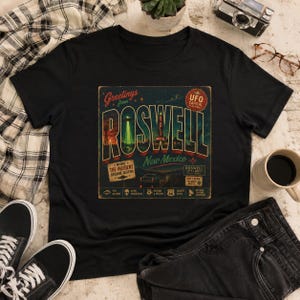 May include: A black short-sleeve t-shirt featuring a vintage-style graphic print that reads Greetings from ROSWELL New Mexico. The design includes a UFO, a car, desert landscapes, and text labels such as UFO Capital of the World and Starlit Motel.