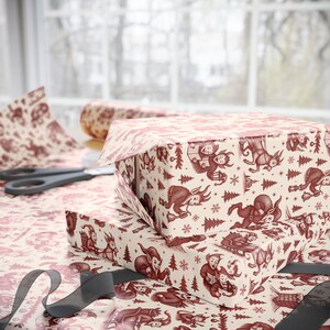 May include: Two gift boxes wrapped in cream-colored paper with a repeating pattern of red illustrations of elves, trees, and snowflakes. A roll of matching wrapping paper and scissors are visible in the background. A gray ribbon is in the foreground.