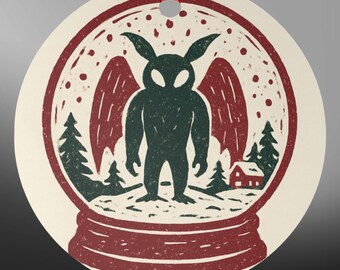 May include: A round, cream-colored ornament featuring a red and dark green graphic print of a winged creature inside a snow globe. The design includes stylized pine trees, a small house, and falling snow, finished with a red ribbon for hanging.