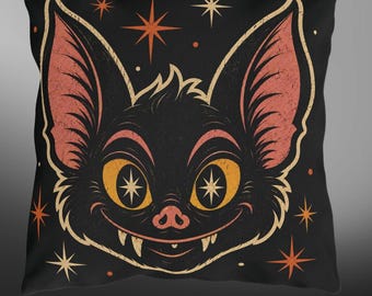 May include: A square throw pillow with a black background featuring a centered illustration of a bat face. The bat has large orange eyes with star-shaped pupils, pink inner ears, and visible fangs. The design includes orange and cream star accents.