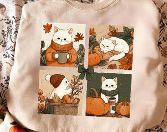 May include: A beige crewneck sweatshirt featuring a four-panel graphic print of white cats in autumn settings. The illustrations include cats with pumpkins, fall leaves, knitted sweaters, hats, scarves, and mugs, all centered with a small green bow.