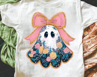 May include: A white short-sleeve t-shirt featuring a central graphic of a ghost adorned with a large pink glittery bow. The ghost is decorated with a floral pattern of pink and yellow roses, dark green leaves, and blue accents.