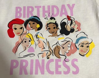 May include: White t-shirt featuring the text "BIRTHDAY PRINCESS" in pink, with illustrations of various princess characters. The shirt has a crew neck and is made of a soft material.