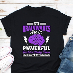 May include: Black t-shirt with a purple brain graphic and the text "MY BRAINWAVES Are So POWERFUL Doctors Study Them EPILEPSY AWARENESS". The shirt also features a purple ribbon and lightning bolts.