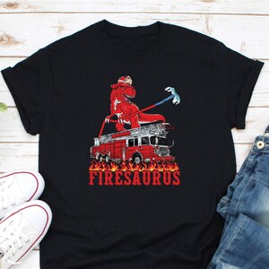 Op de afbeelding: Zwart T-shirt met een rode dinosaurus brandweerman op een rode brandweerwagen. De tekst "FIRESASAURUS" is rood. De dinosaurus draagt een brandweerhelm en houdt een gereedschap vast. De brandweerwagen is omringd door vlammen.